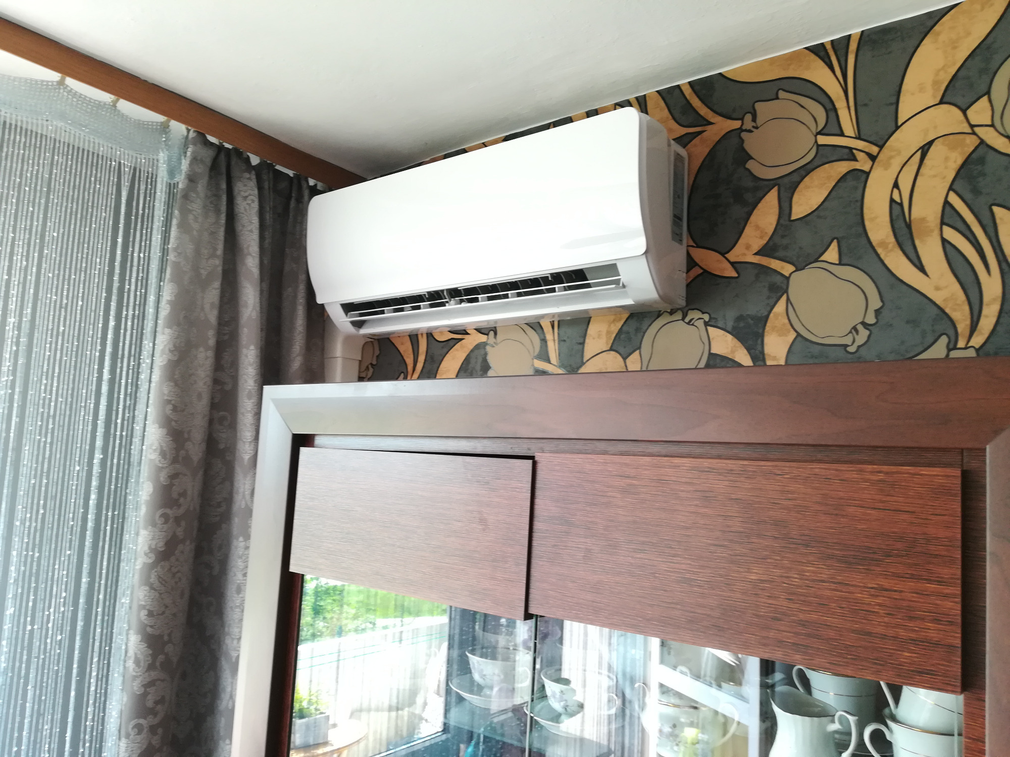 Biały klimatyzator ścienny zamontowany nad drewnianą witryną z przeszklonymi drzwiami, na tle ściany z wzorzystą tapetą w odcieniach szarości i złota.