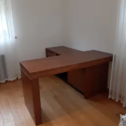 Kątowy, drewniany blat biurka w jasnym pomieszczeniu z drewnianą podłogą, oświetlonym przez okno z białymi, zwiewnymi zasłonami.