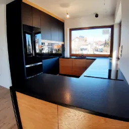 Nowoczesna kuchnia z czarnymi szafkami i drewnianymi frontami, czarny blat, okno z widokiem na domy jednorodzinne, minimalistyczne oświetlenie.