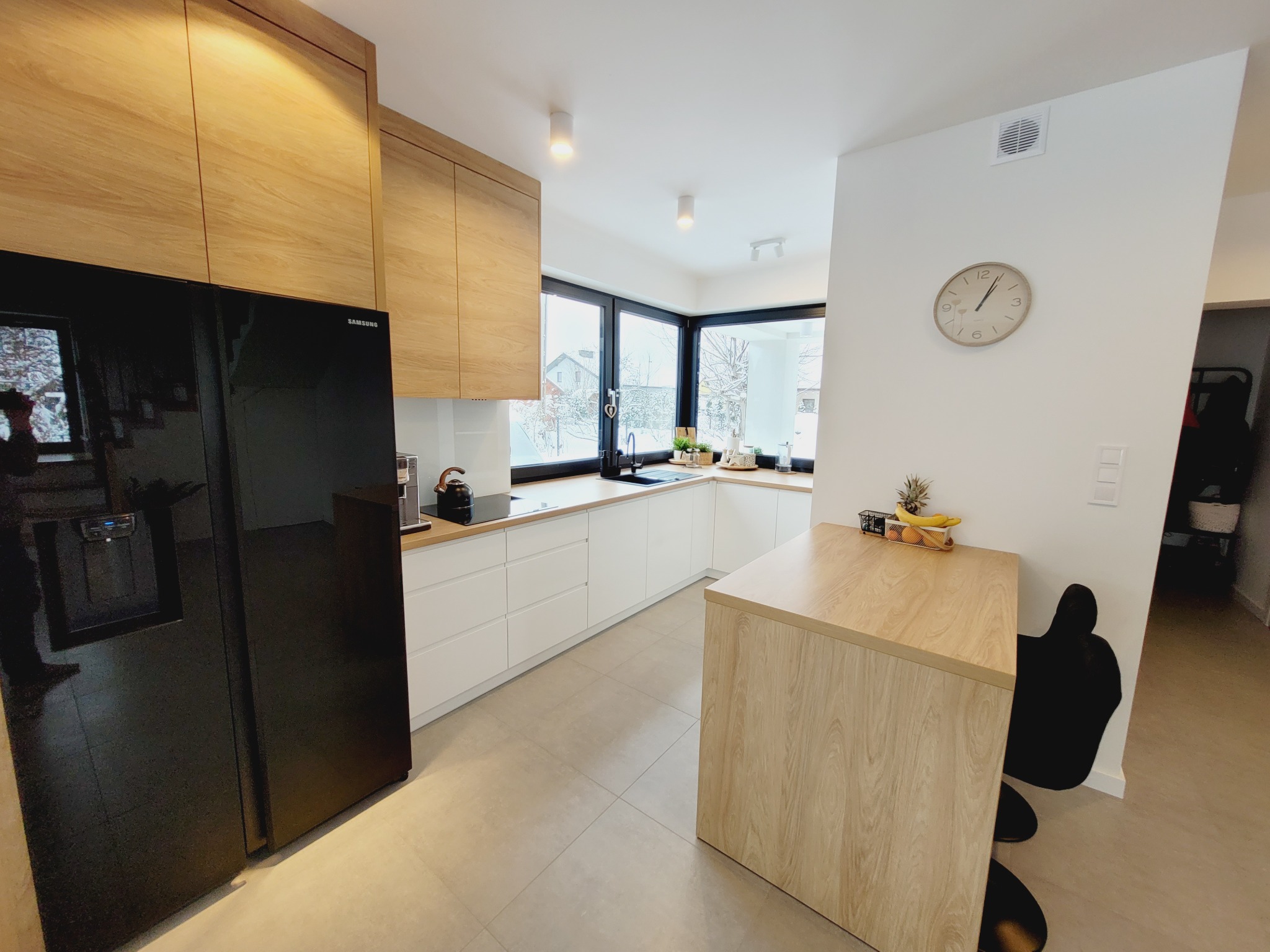 Nowoczesna, jasna kuchnia z białymi szafkami i drewnianymi elementami, widok na zimowy krajobraz przez narożne okno, czarna lodówka Samsung po lewej stronie, zegar na ścianie.