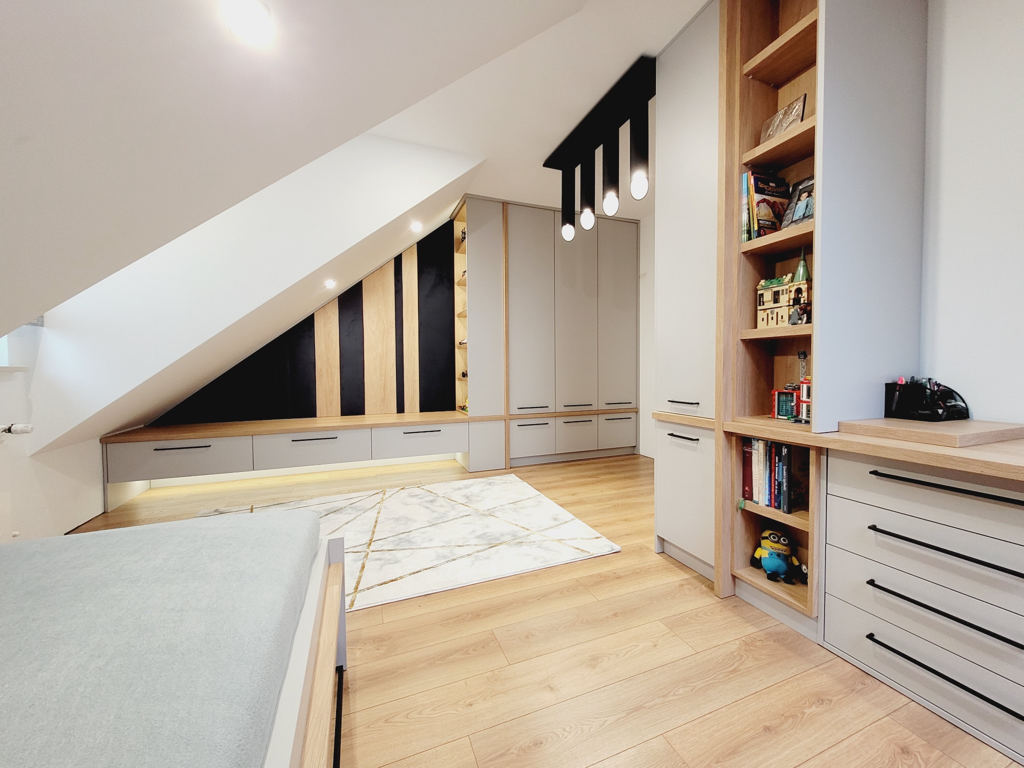 Adaptacja poddasza ze skośnym sufitem: zabudowa meblowa z szarymi szafkami i drewnianymi akcentami, oświetlenie punktowe, regał z książkami i zabawkami, dywan w geometryczne wzory na drewnianej...