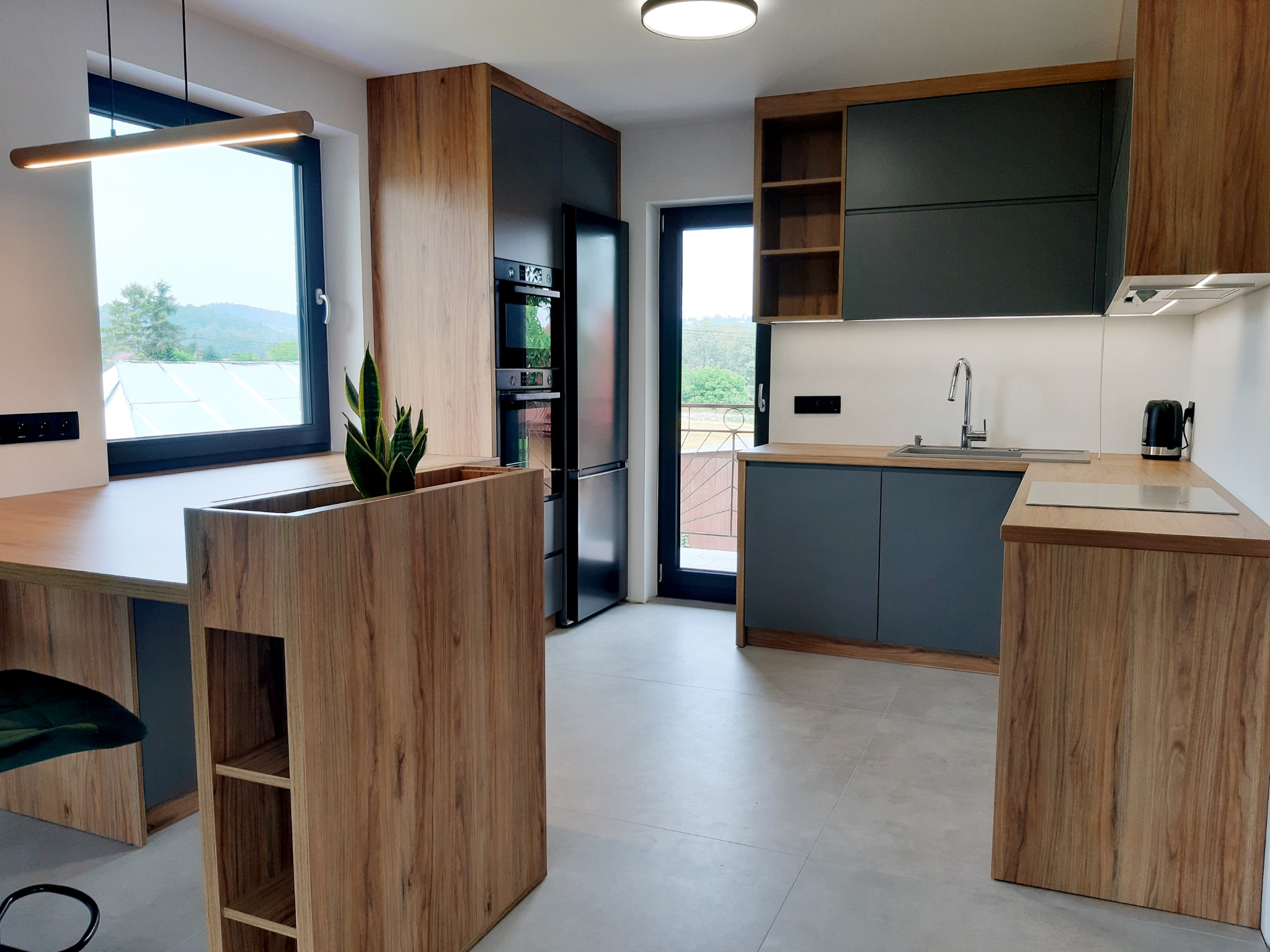 Nowoczesna kuchnia z drewnianymi i grafitowymi szafkami, wyspą z wbudowaną donicą i widokiem na góry za oknem.
