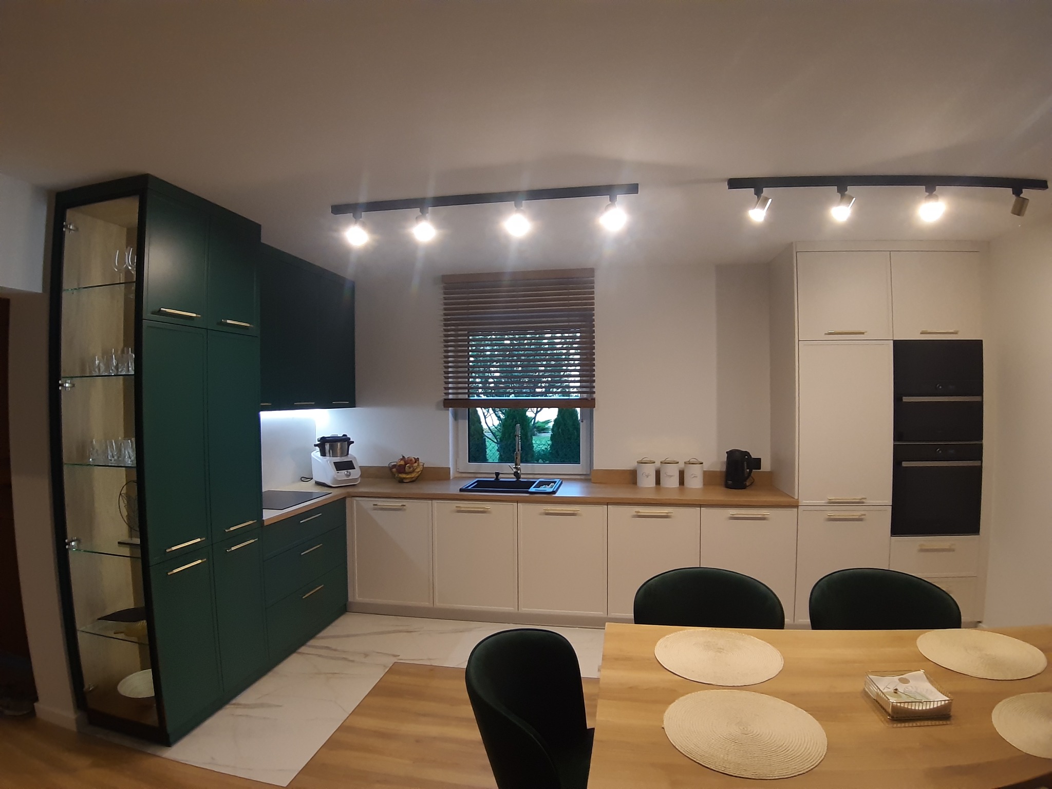 Nowoczesna kuchnia z zielonymi szafkami, białymi frontami i drewnianym blatem, widok na stół z zielonymi krzesłami i okno z żaluzjami.