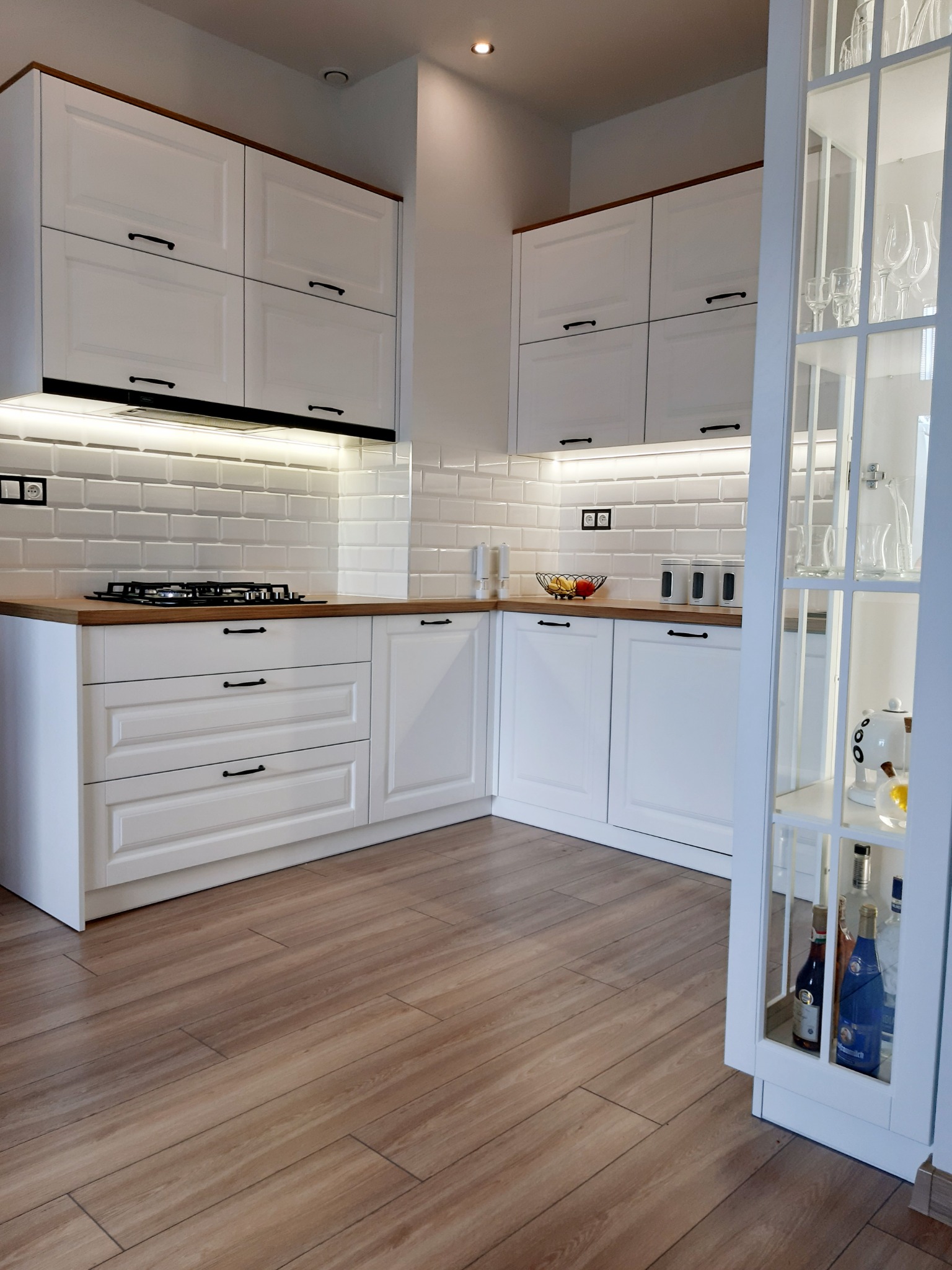 Jasna kuchnia z białymi, frezowanymi frontami szafek, drewnianym blatem i białymi płytkami na ścianie, uzupełniona o przeszkloną witrynę z dekoracyjnym oświetleniem i butelkami.