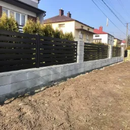 Nowoczesne ogrodzenie z poziomych, czarnych paneli zamontowanych na betonowej podmurówce i słupkach, widoczne na tle domów w słoneczny dzień.