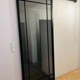 G-Weld - Szklane drzwi przesuwne w czarnej metalowej ramie, widoczne na tle białej ściany i parkietu ułożonego w jodełkę.