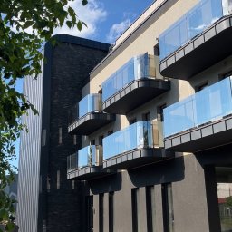 G-Weld - Nowoczesny budynek wielorodzinny z balkonami zabezpieczonymi przezroczystymi taflami szkła, widok z poziomu chodnika z fragmentem drzewa po lewej stronie i błękitnym niebem z chmurami w tle.