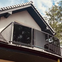 Balkon z czarną, szklaną balustradą i metalowymi słupkami, widok z dołu na tle błękitnego nieba i korony drzewa.