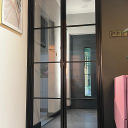 G-Weld - Dwuskrzydłowe drzwi wewnętrzne z czarną metalową ramą i szprosami, widoczne wnętrze z szarą podłogą i ciemnymi drzwiami w tle.