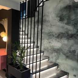 G-Weld - Nowoczesne schody z podświetleniem LED, czarną metalową balustradą i roślinami doniczkowymi, na tle ściany z efektem dekoracyjnym.