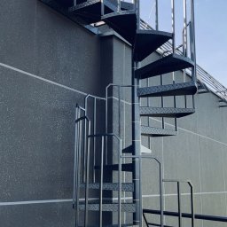 G-Weld - Zewnętrzne czarne schody spiralne z metalu z ażurową balustradą, prowadzące na dach budynku o szarej, chropowatej elewacji, widoczne na tle błękitnego nieba.
