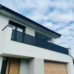 Nowoczesny dom z balkonem zabezpieczonym czarną, pionową balustradą, widok z dołu na białą elewację, drewniane drzwi wejściowe i garażowe, błękitne niebo w tle.