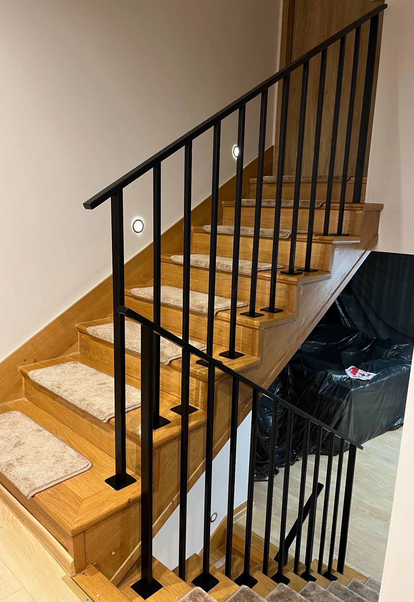 Drewniane schody z czarną, metalową balustradą i beżowymi nakładkami na stopniach, oświetlone punktowymi lampami ściennymi. W tle widoczny mebel przykryty czarną folią.