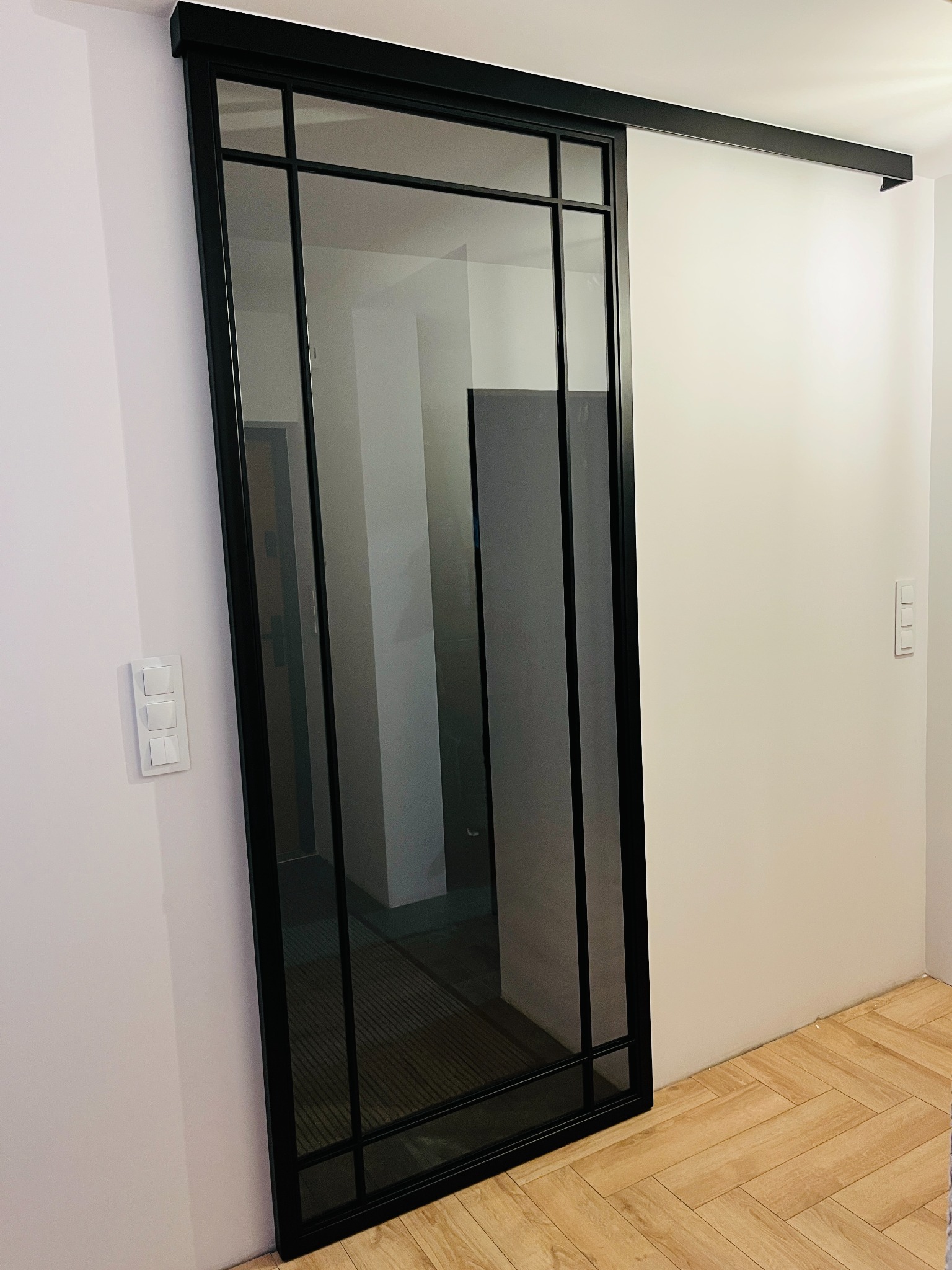 Szklane drzwi przesuwne w czarnej metalowej ramie, widoczne na tle białej ściany i parkietu ułożonego w jodełkę.
