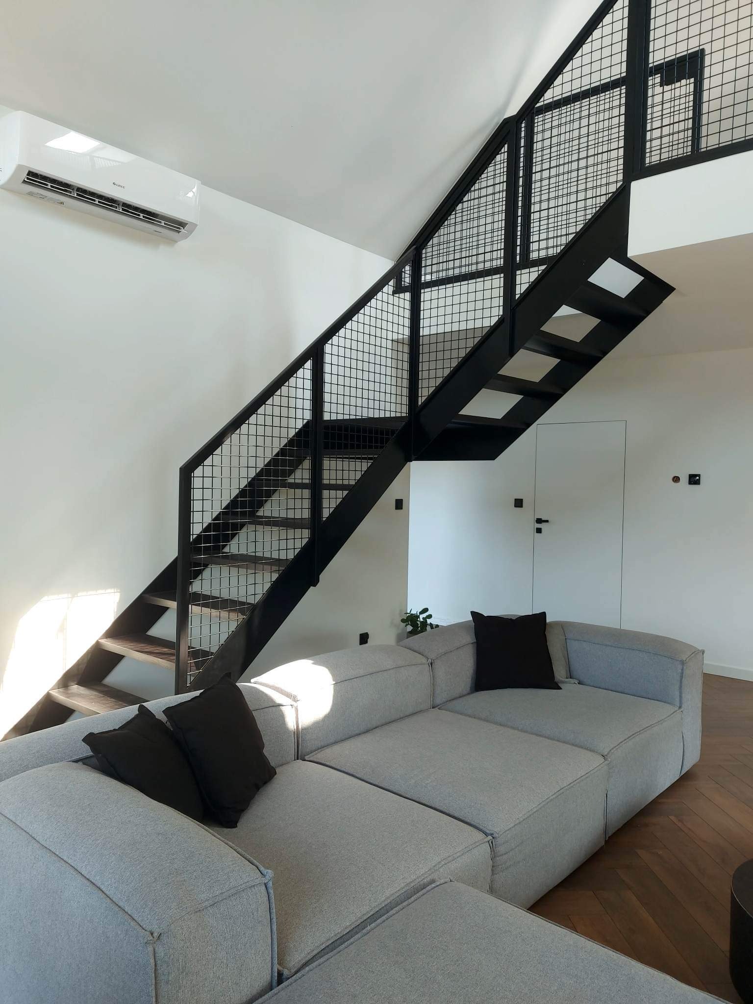 Nowoczesne wnętrze z czarnymi schodami z metalową siatką, drewnianymi stopniami i szarą sofą modułową z czarnymi poduszkami, oświetlone naturalnym światłem.