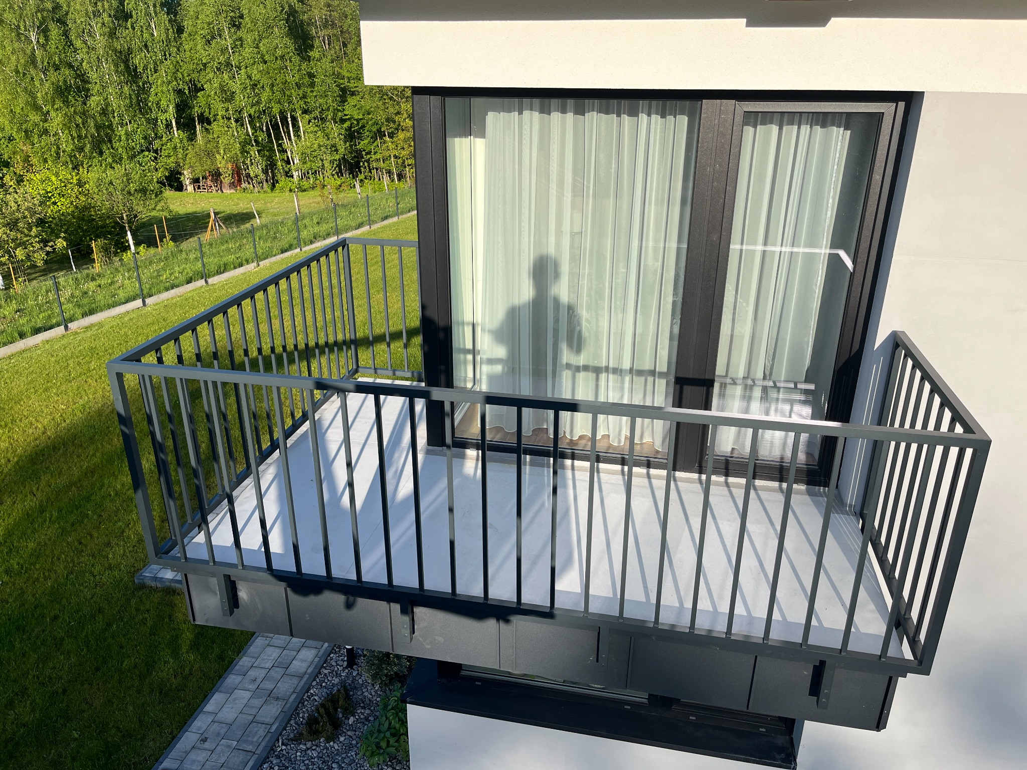 Nowoczesny balkon z czarną metalową balustradą o minimalistycznym designie, widok z góry na tle zielonego trawnika i lasu, cień osoby widoczny na zasłonie w oknie.
