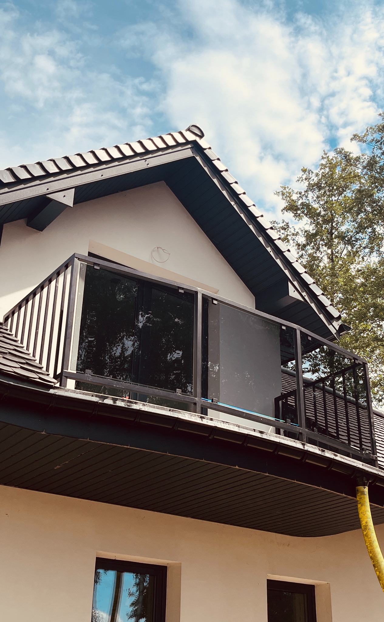 Balkon z czarną, szklaną balustradą i metalowymi słupkami, widok z dołu na tle błękitnego nieba i korony drzewa.