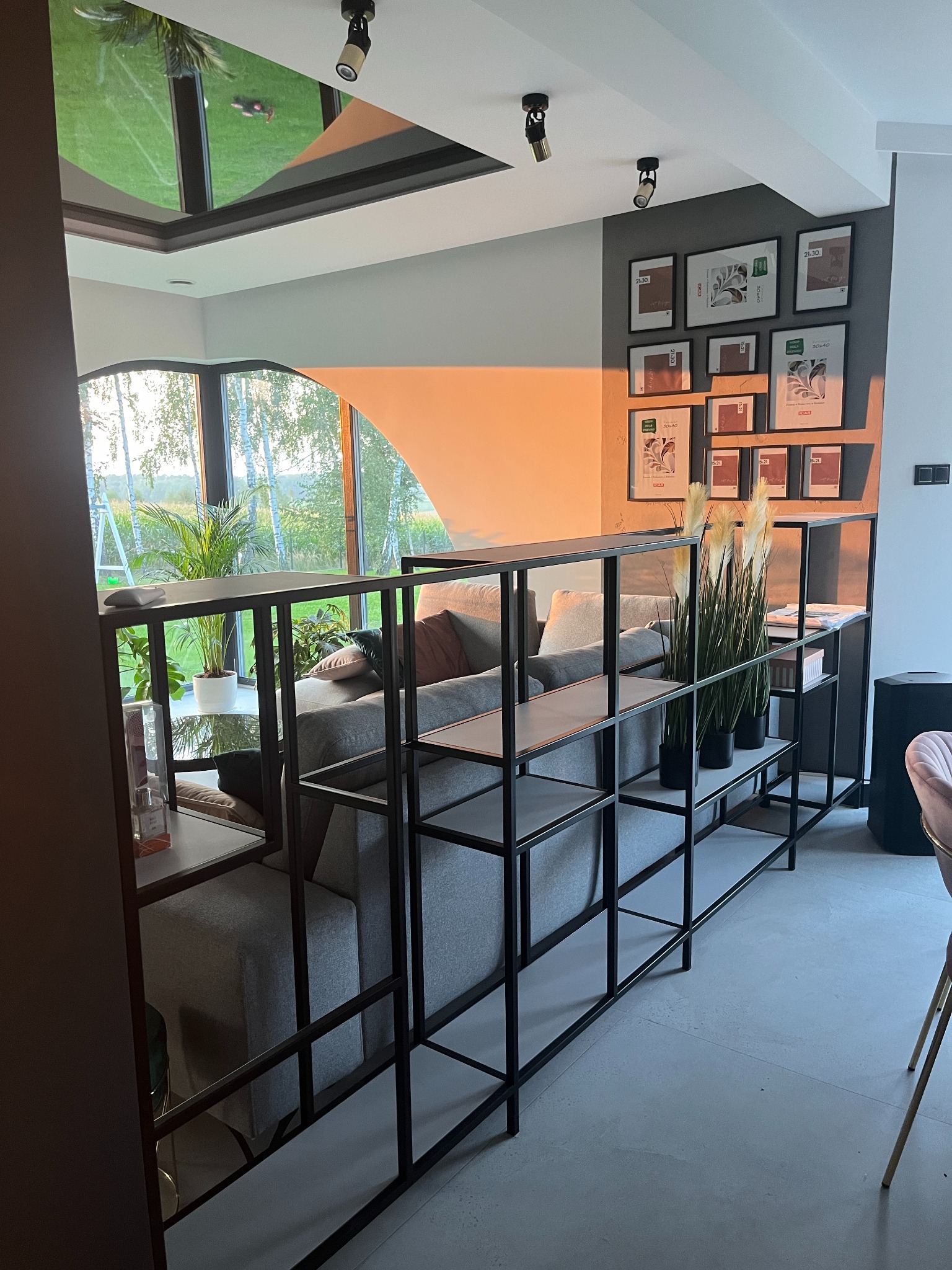 Metalowa konstrukcja regału z półkami, dzieląca przestrzeń salonu, z widoczną sofą w tle i oknem z widokiem na zieleń, oświetlona popołudniowym światłem.