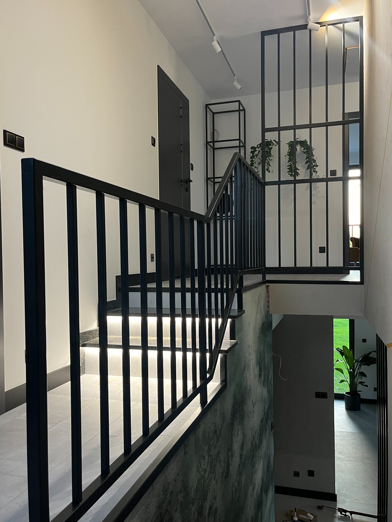 Nowoczesne schody z podświetlanymi stopniami i czarną, minimalistyczną balustradą, widok z góry na dół, z elementami dekoracyjnymi jak rośliny wiszące i półka w tle.