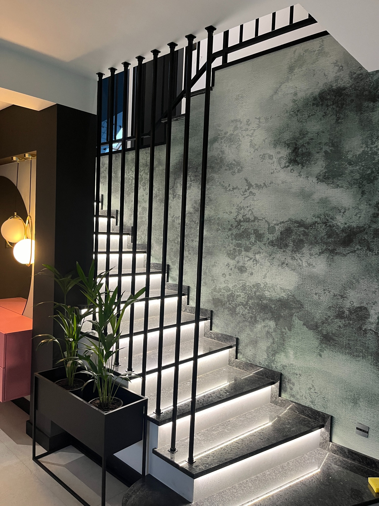 Nowoczesne schody z podświetleniem LED, czarną metalową balustradą i roślinami doniczkowymi, na tle ściany z efektem dekoracyjnym.