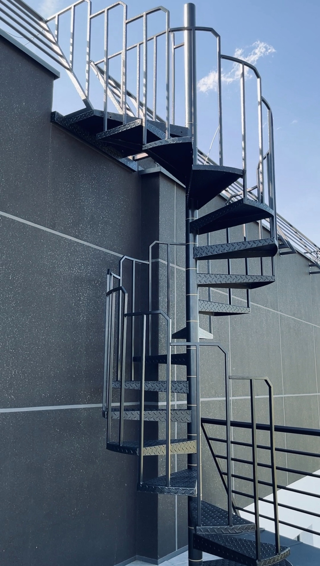 Zewnętrzne czarne schody spiralne z metalu z ażurową balustradą, prowadzące na dach budynku o szarej, chropowatej elewacji, widoczne na tle błękitnego nieba.