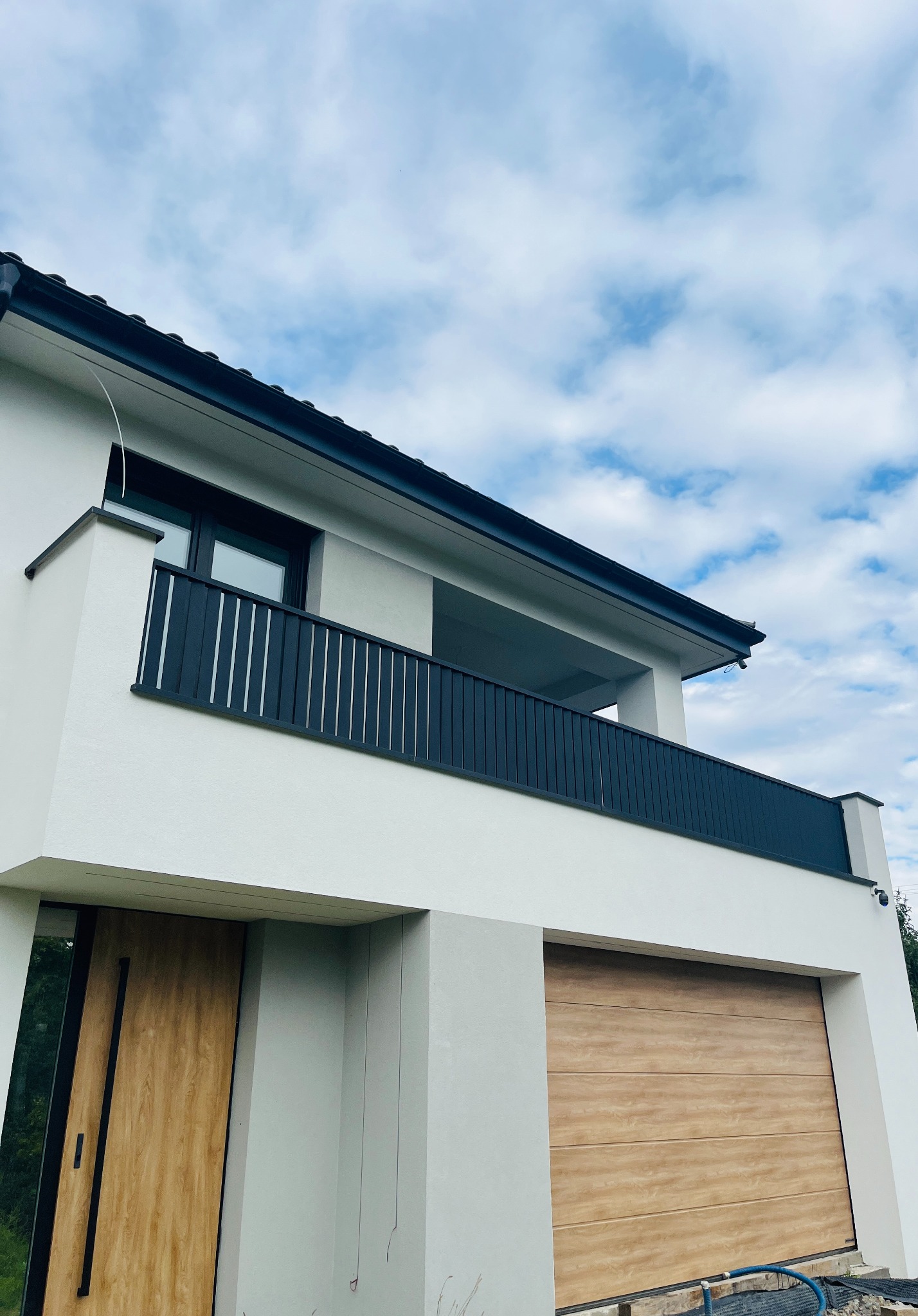 Nowoczesny dom z balkonem zabezpieczonym czarną, pionową balustradą, widok z dołu na białą elewację, drewniane drzwi wejściowe i garażowe, błękitne niebo w tle.