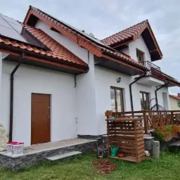 Biały dom jednorodzinny z brązowymi elementami wykończeniowymi, panelami słonecznymi na dachu i drewnianym tarasem, widok z boku na zadbany trawnik i elementy wyposażenia ogrodowego.