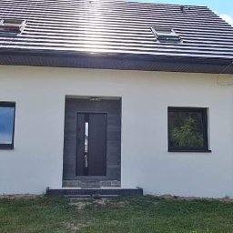 Dom-Wit - Biały, ocieplony dom jednorodzinny z ciemną stolarką okienną i drzwiową oraz grafitowym dachem z lukarnami, widok z przodu na tle zielonego trawnika i zabudowy sąsiedzkiej.