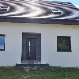 Biały, ocieplony dom jednorodzinny z ciemną stolarką okienną i drzwiową oraz grafitowym dachem z lukarnami, widok z przodu na tle zielonego trawnika i zabudowy sąsiedzkiej.