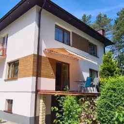 Dwupiętrowy dom z białą elewacją, drewnianymi panelami i balkonem z czerwoną barierką, otoczony zielenią drzew i krzewów w słoneczny dzień.