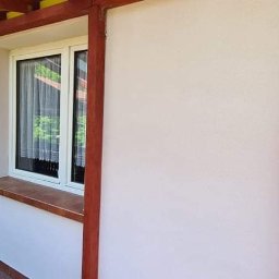Dom-Wit - Ściana budynku po nałożeniu jasnego tynku strukturalnego, z widoczną ramą okienną i elementami drewnianymi konstrukcji werandy.