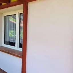 Ściana budynku po nałożeniu jasnego tynku strukturalnego, z widoczną ramą okienną i elementami drewnianymi konstrukcji werandy.