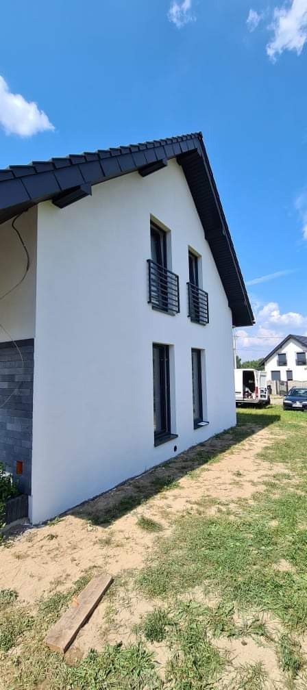 Dwukondygnacyjny dom z białą elewacją i czarnym dachem, widok z boku na tle błękitnego nieba z chmurami, z widocznymi balustradami balkonowymi i fragmentem wykończenia z szarej cegły.