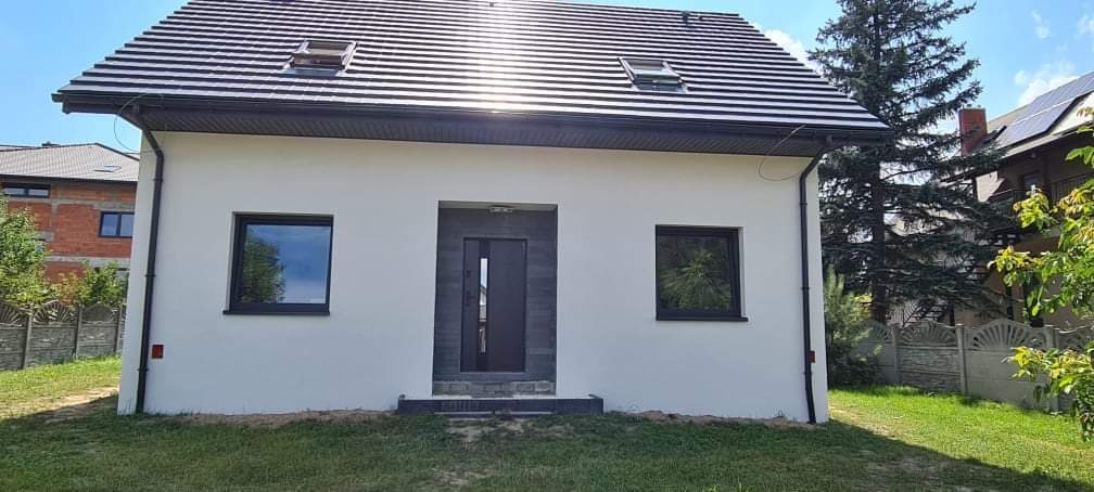 Biały, ocieplony dom jednorodzinny z ciemną stolarką okienną i drzwiową oraz grafitowym dachem z lukarnami, widok z przodu na tle zielonego trawnika i zabudowy sąsiedzkiej.