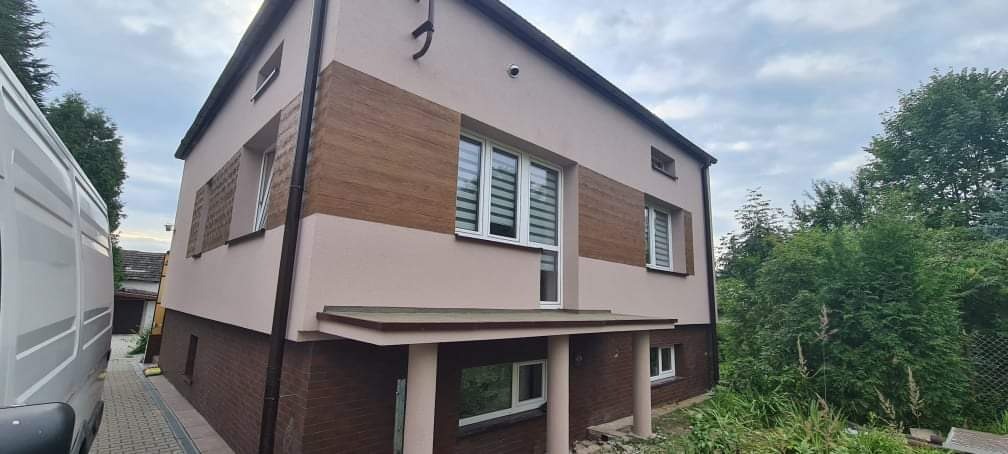 Dwukondygnacyjny dom z elewacją w kolorze beżowym i brązowym, z poziomymi pasami imitującymi drewno, widok z boku z zaparkowanym białym vanem.