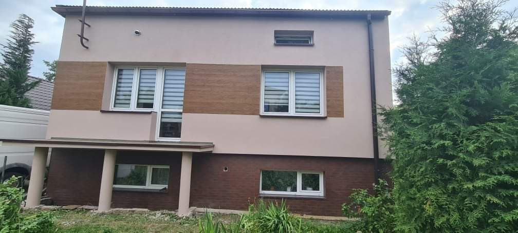 Dwukondygnacyjny dom z elewacją w odcieniach beżu i brązu, z elementami imitującymi drewno na górnej kondygnacji i cegłę na dolnej, z białymi ramami okiennymi i szarymi roletami poziomymi, widok...