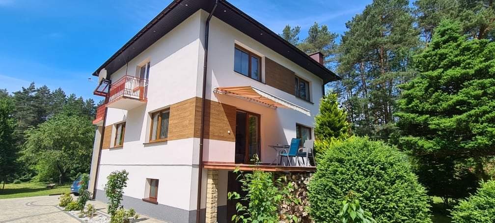 Dwupiętrowy dom z białą elewacją, drewnianymi panelami i balkonem z czerwoną barierką, otoczony zielenią drzew i krzewów w słoneczny dzień.