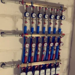 Rozdzielacz instalacji wodnej z licznymi wodomierzami, rury w izolacji niebieskiej i czerwonej, zamontowany na ścianie z betonu komórkowego.