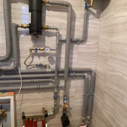 Złożona instalacja grzewcza z rurami w izolacji, czarnym filtrem, manometrem i rozdzielaczem podłogówki, zamontowana na ścianie wyłożonej płytkami imitującymi drewno.