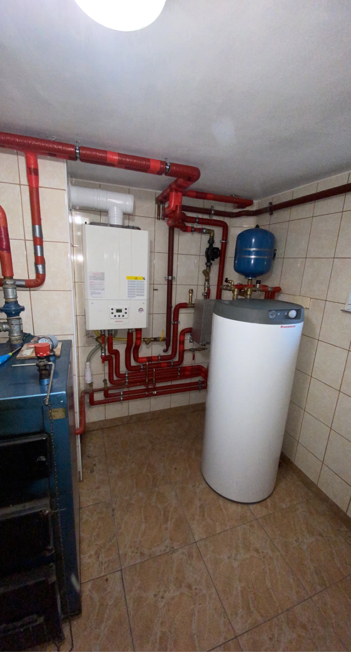 Wnętrze kotłowni z piecem gazowym, zasobnikiem ciepłej wody i rozbudowaną instalacją grzewczą z rur w izolacji termicznej, widoczne płytki ceramiczne na ścianach i podłodze.