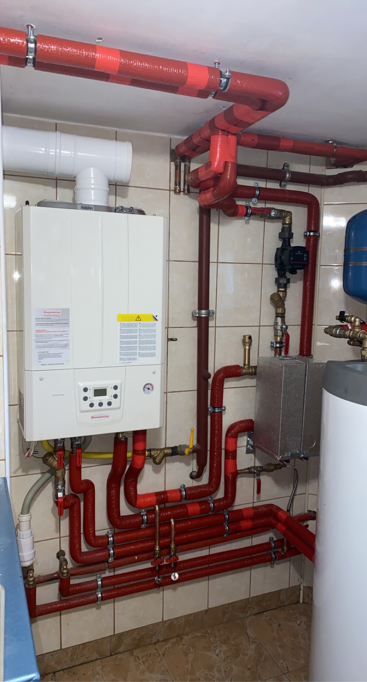 Nowoczesny piec gazowy Immergas z licznymi czerwonymi rurami instalacji grzewczej, częściowo zaizolowanymi, zamontowany na ścianie z beżowych płytek.