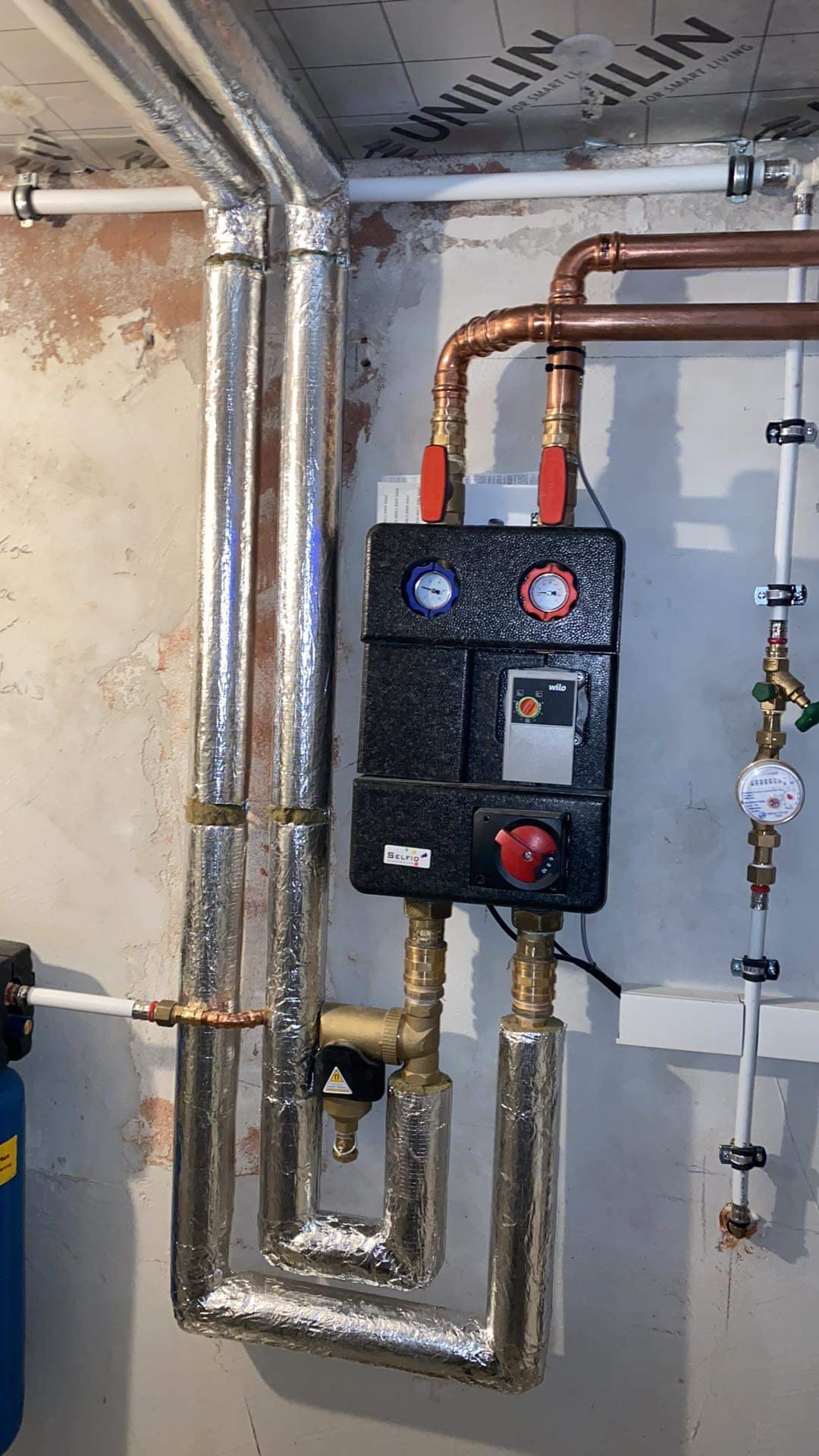 Instalacja grzewcza z czarną skrzynką rozdzielczą, miedzianymi rurami i zaworami z czerwonymi pokrętłami, rury izolowane folią aluminiową, widoczny wodomierz i filtr wody.