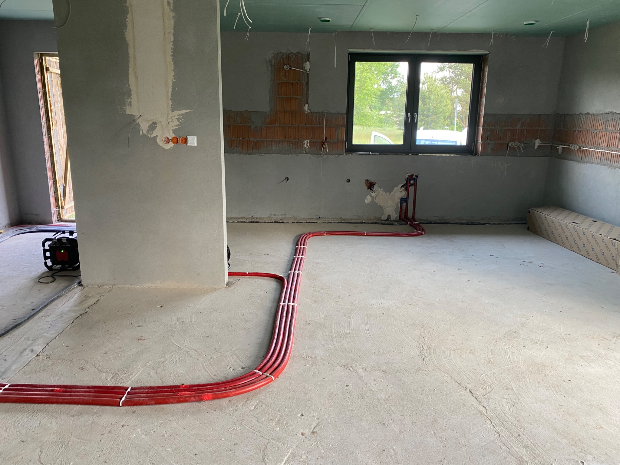 Wnętrze w trakcie prac wykończeniowych z widocznymi czerwonymi rurami instalacji grzewczej ułożonymi na surowym betonowym podłożu, ściany w stanie deweloperskim, okno z widokiem na zieleń.