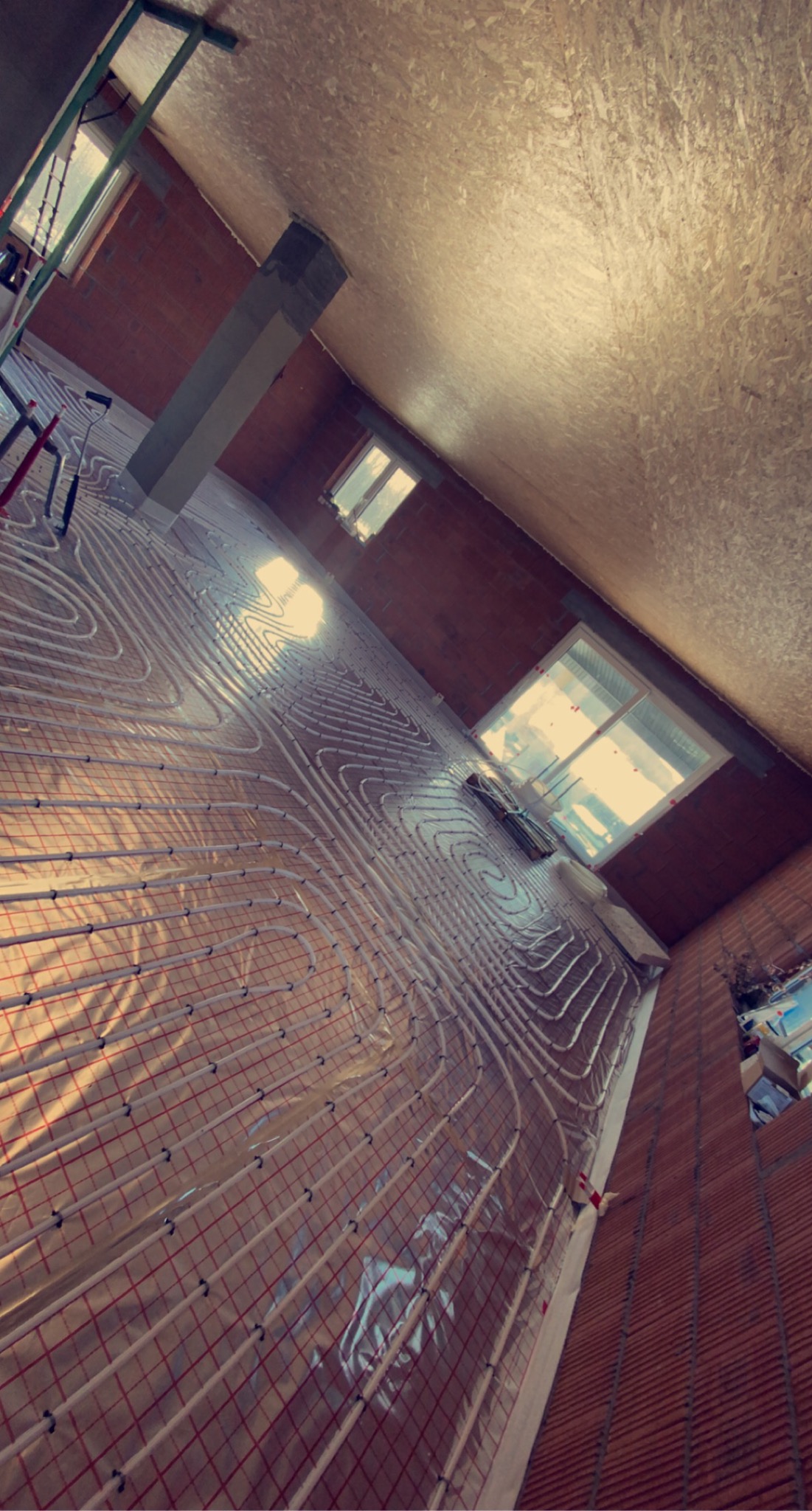 Widok z góry na rozłożone rury ogrzewania podłogowego na folii z czerwoną siatką, w surowym wnętrzu z oknami i ceglanymi ścianami, przygotowane do wylewki.