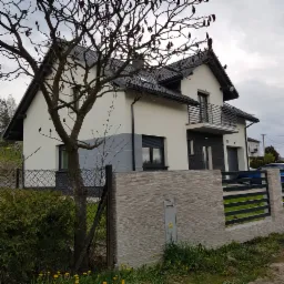 Nowoczesny dom jednorodzinny z balkonem, wykończony w bieli i szarości, z widocznym fragmentem ogrodzenia z elementami imitującymi kamień i drzewem z bordowymi pąkami na pierwszym planie.