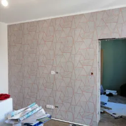 Ściana po tapetowaniu z geometrycznym wzorem w odcieniach szarości i różu, widoczne rolki tapety i gniazdka elektryczne, w tle otwarte drzwi do innego pomieszczenia.