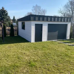 Nowoczesny, biały garaż z szarą bramą segmentową i dachem z blachy trapezowej, widziany z perspektywy trawnika w słoneczny dzień.