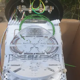 Otwarta, czarna studzienka telekomunikacyjna z widocznym wnętrzem: biała kaseta światłowodowa z kolorowymi włóknami, metalowe złącza i kable zasilające. Na kasecie widoczne odręczne napisy.