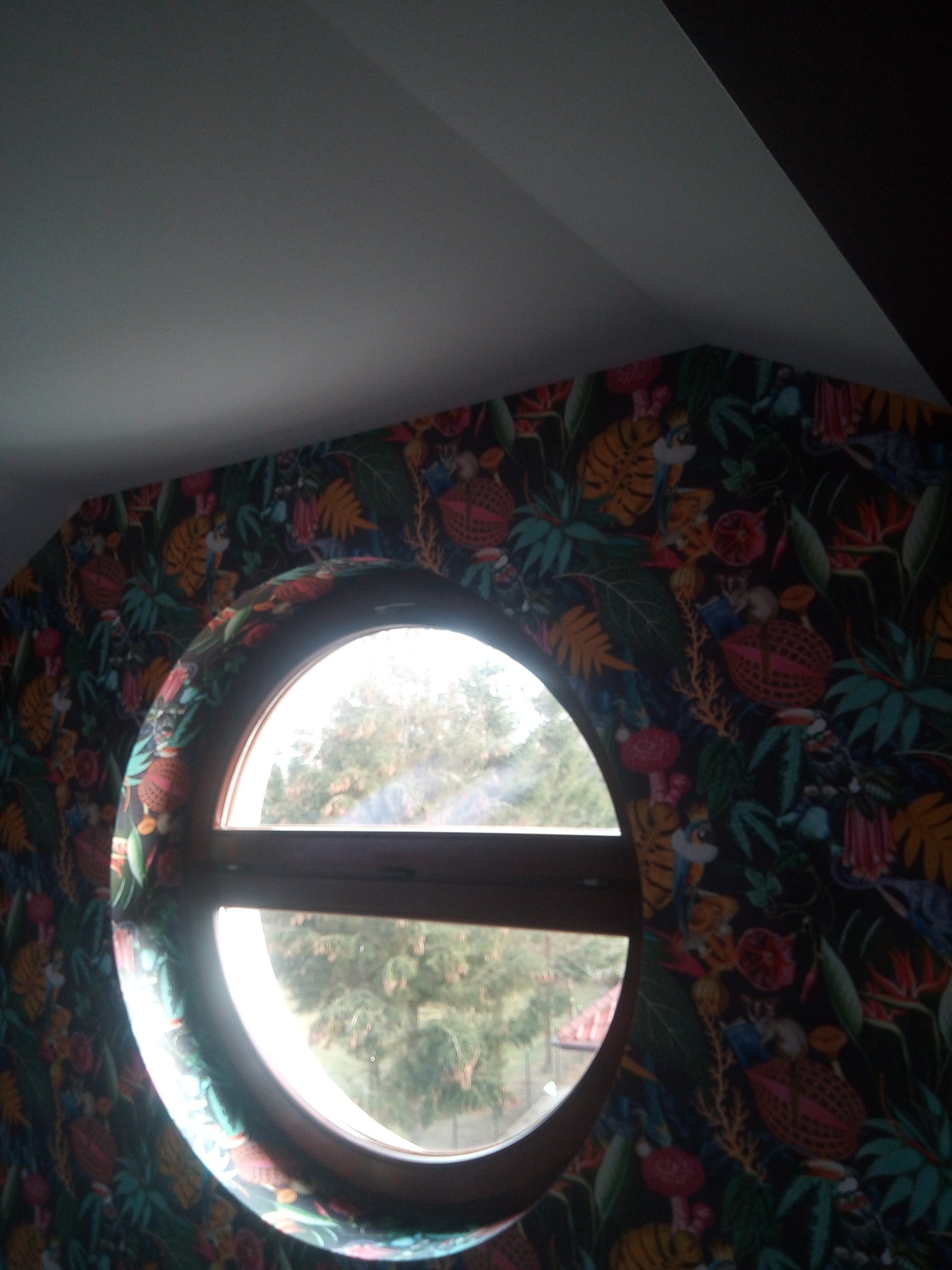 Okrągłe okno z widokiem na drzewo, otoczone tapetą w motywy dżungli z tygrysami, papugami i kwiatami, na poddaszu.