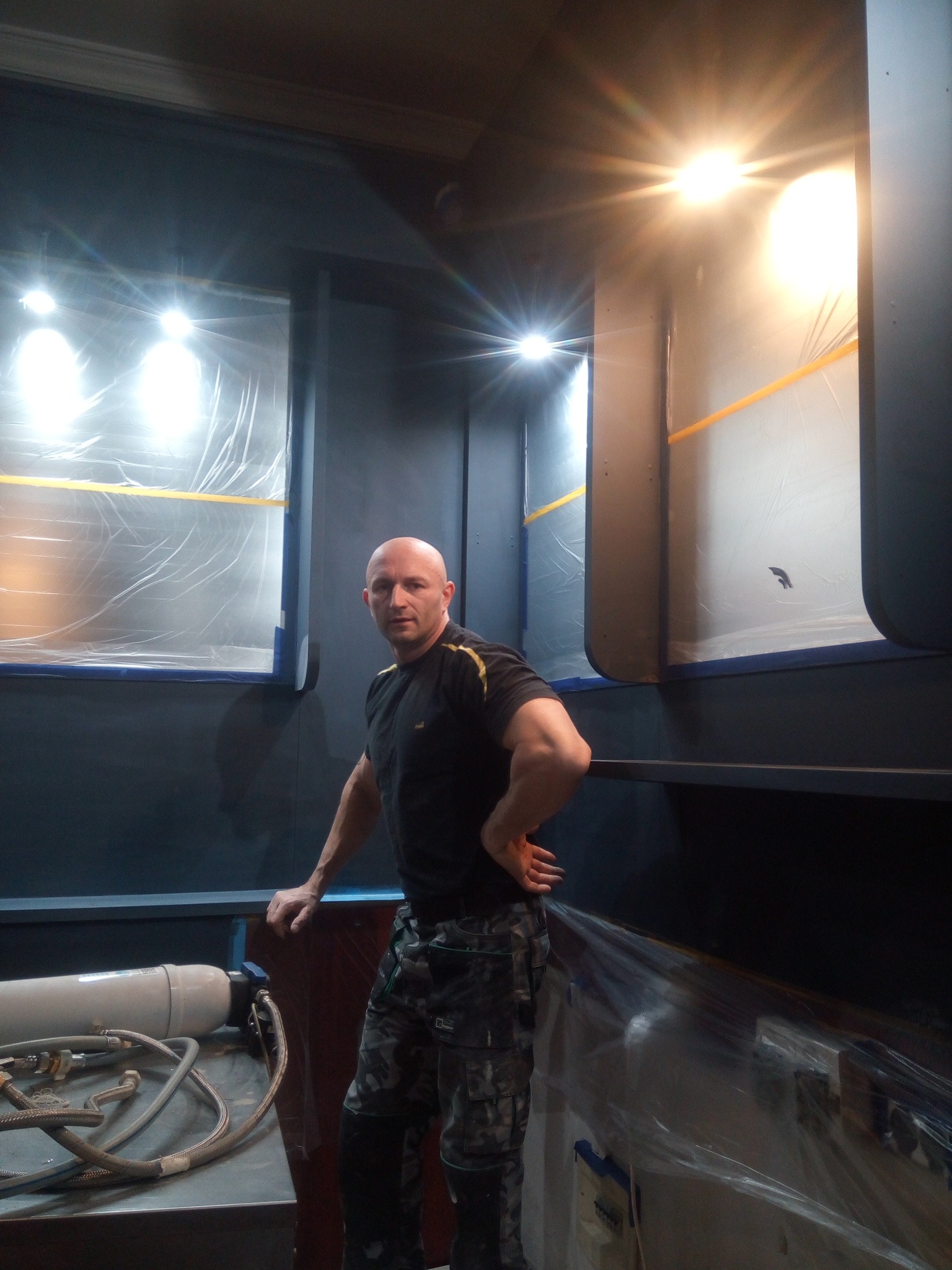 Mężczyzna w trakcie malowania wnętrza na ciemny kolor, okna zabezpieczone folią malarską, widoczny sprzęt malarski.